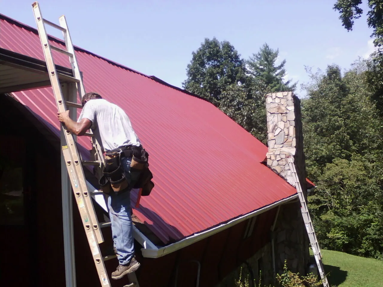 Roofer on a ladder beside a red metal roof and stone chimney — Soffit & Fascia Repair by Roofing Brothers in Conway Springs
